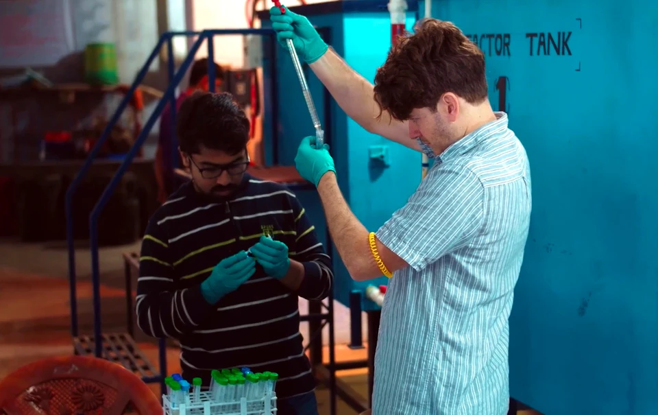 Case van Genuchten is dropping a liquid into a test tube in front of big blue metal tanks. A colleague is standing next to van Genuchten and looking at some test tubes. They are both wearing gloves.