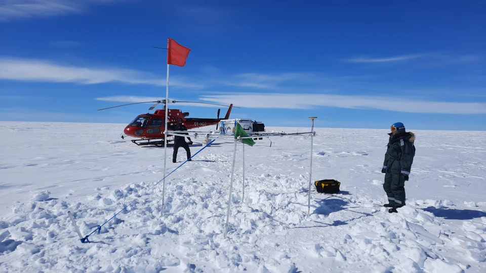 En målestation på Indlandsisen. Hvis grund, to mennesker iført overtøj kigger på stationen. Begved er der en rød helikopter.