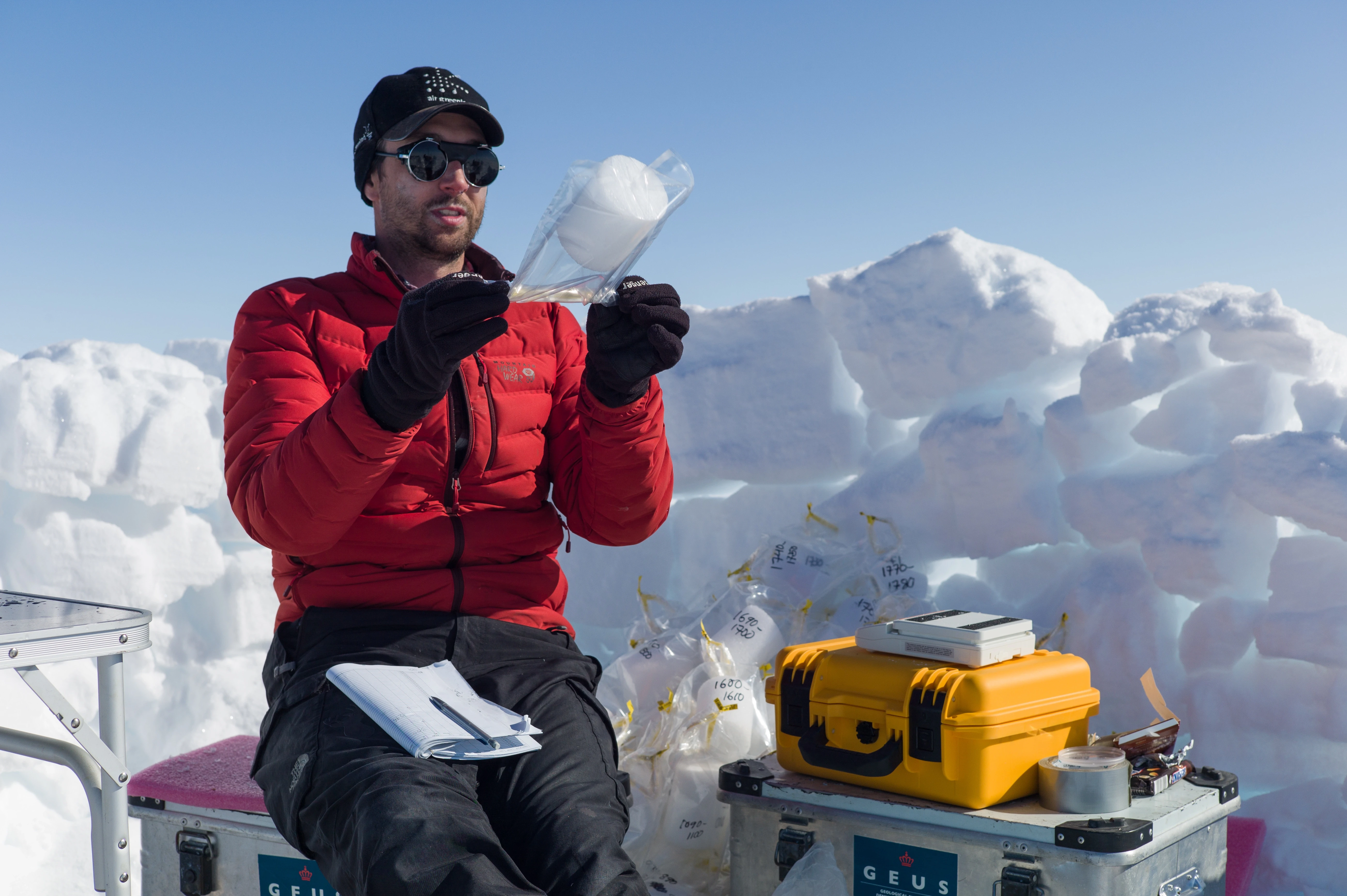 A person is sitting in a digged out area of ice putting small cores of ice in bags.