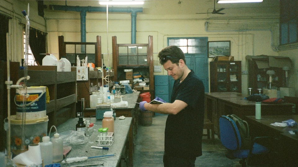 Et menneske står i et laboratorium. Personen skriver i en bog. Der er laboratorieudstyr foran personen på et bord.