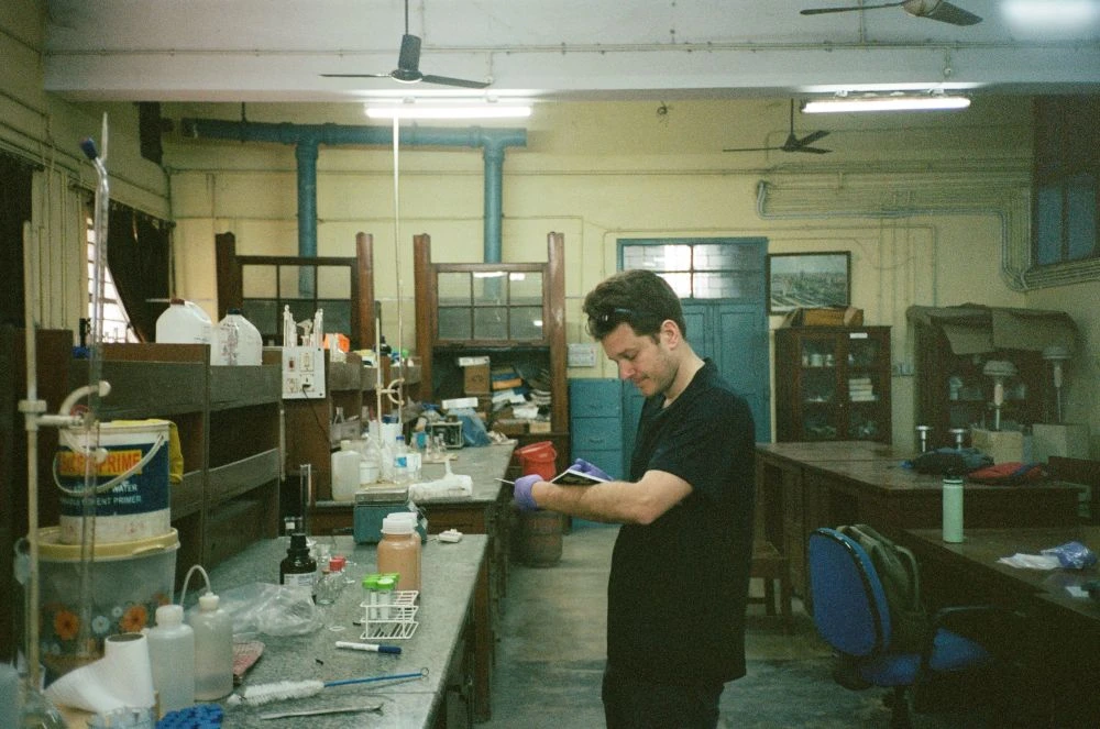 Et menneske står i et laboratorium. Personen skriver i en bog. Der er laboratorieudstyr foran personen på et bord.