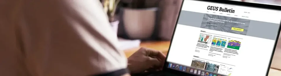 A picture taken over the shoulder of a person shows a screen in focus with the GEUS Bulletin website on it.