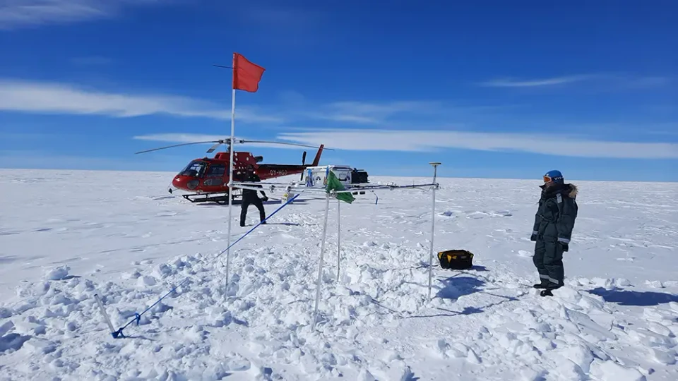En målestation på Indlandsisen. Hvis grund, to mennesker iført overtøj kigger på stationen. Begved er der en rød helikopter.