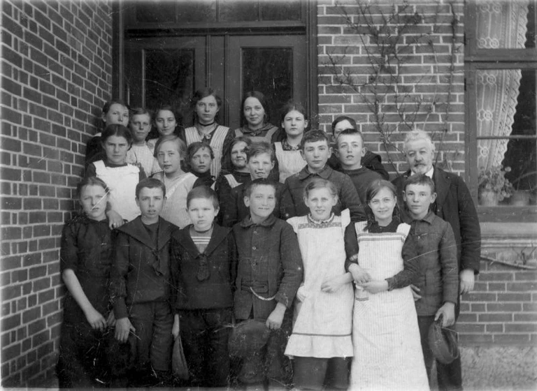 Det ser ud som om at nogle af eleverne fra Ejlstrup Skole smiler. Mon de har fået læst vittighederne fra Dansk Børnetidende højt af lærer Petersen (th)? Billedet er fra 1910.