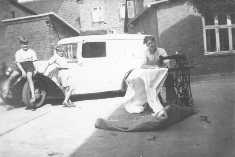 Sadelmager Sven Aage Terkelsen ved symaskinen på gårdspladsen med to drenge som tilskuere op ad firmabilen, en flot Citroen.