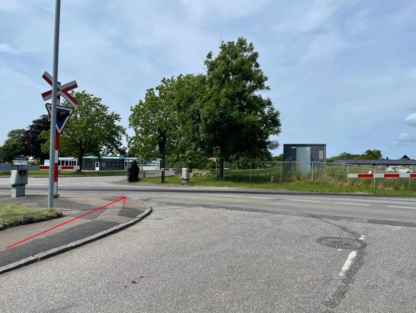 Stor åbning imellem bomme ved blik fra Birkevej mod Sorøvej, den røde pil viser fodgængerens rute fra fortov og ud på Sorøvej og jernbanesporet. Sti bom og vejsignal ses i billedets venstre side. 