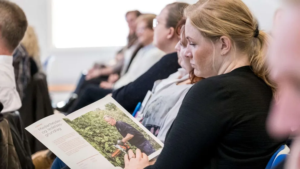 Kvinde læser en A-4 publikation fra stolerække med mange andre deltagere omkring hende